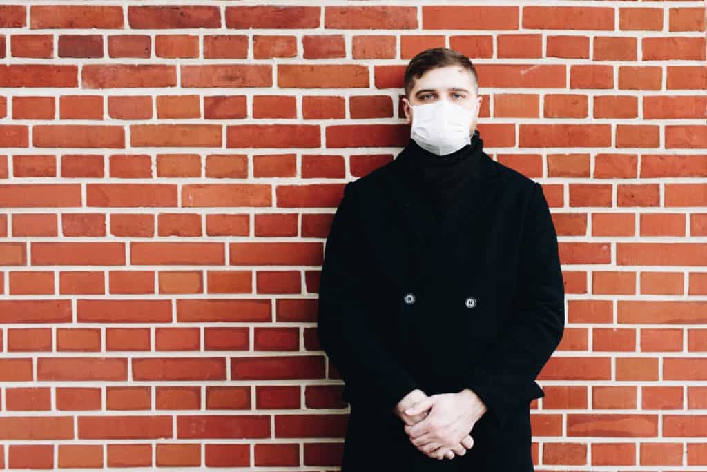 Young caucasian man wearing a surgical mask to protect from coronavirus covid-19 during global pandemic standing against a wall